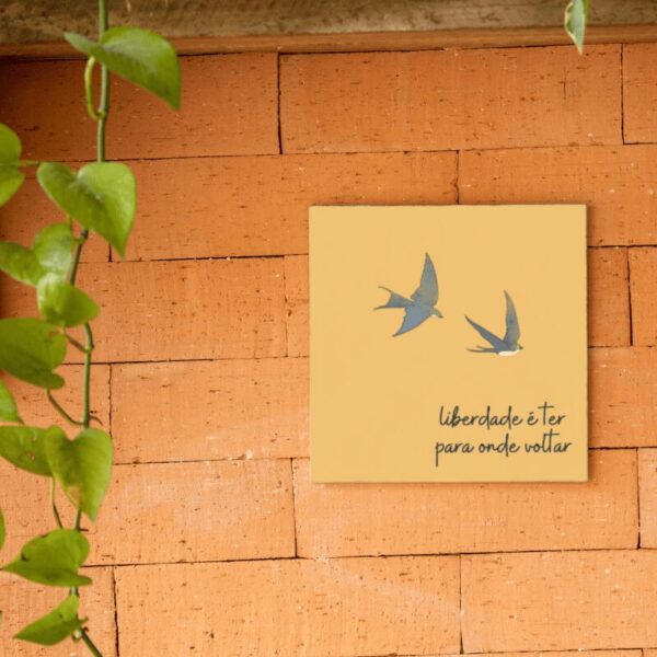 Azulejo amarelo Liberdade é Ter para Onde Voltar com duas aves em voo, pendurado na parede de tijolos