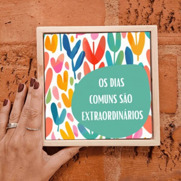 Mão segurando o azulejo Os Dias Comuns São Extraordinários com folhas coloridas e moldura de madeira
