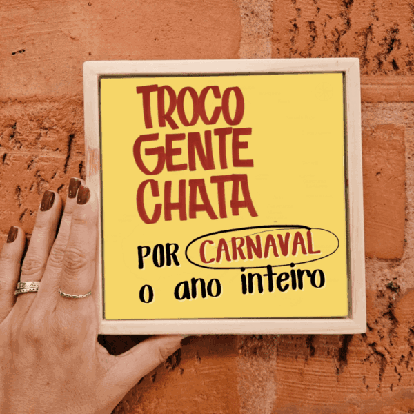 Mão segurando o azulejo amarelo Troco Gente Chata por Carnaval o Ano Inteiro em moldura de madeira